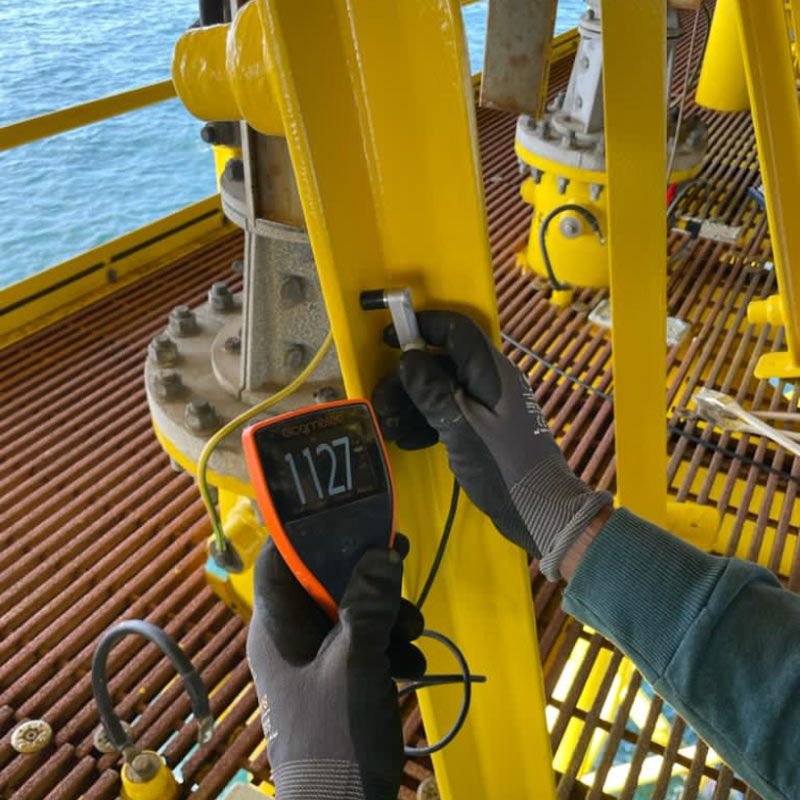 Technician measuring coating thickness on offshore platform steelwork with a digital gauge.