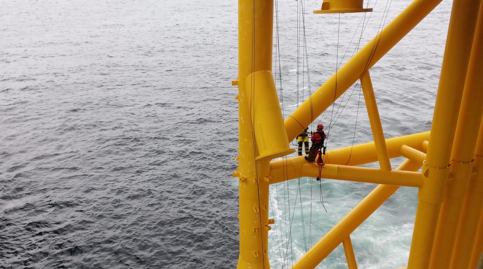 Rope access technicians on a yellow offshore jacket structure above the sea.