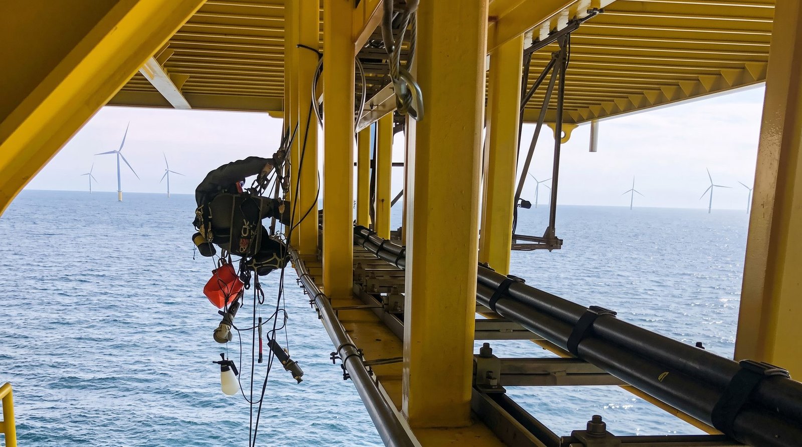 Rope access technician working beneath an offshore platform above the sea, with a wind farm visible in the background.