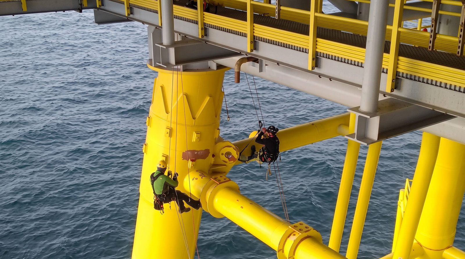Rope access technicians working on a steel offshore foundation structure beneath a platform.