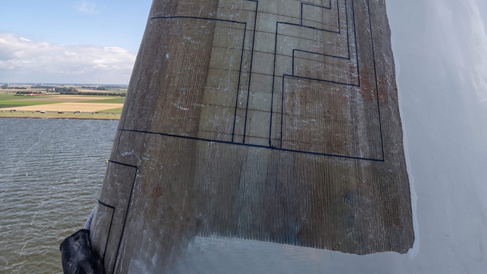 Close-up of a wind turbine rotor blade surface showing marked laminate repair boundaries, with a wind farm landscape in the background.