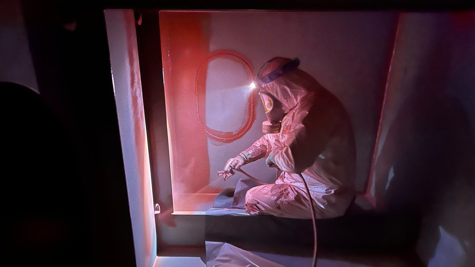 Technician in a protective suit performing surface preparation work inside a steel compartment.