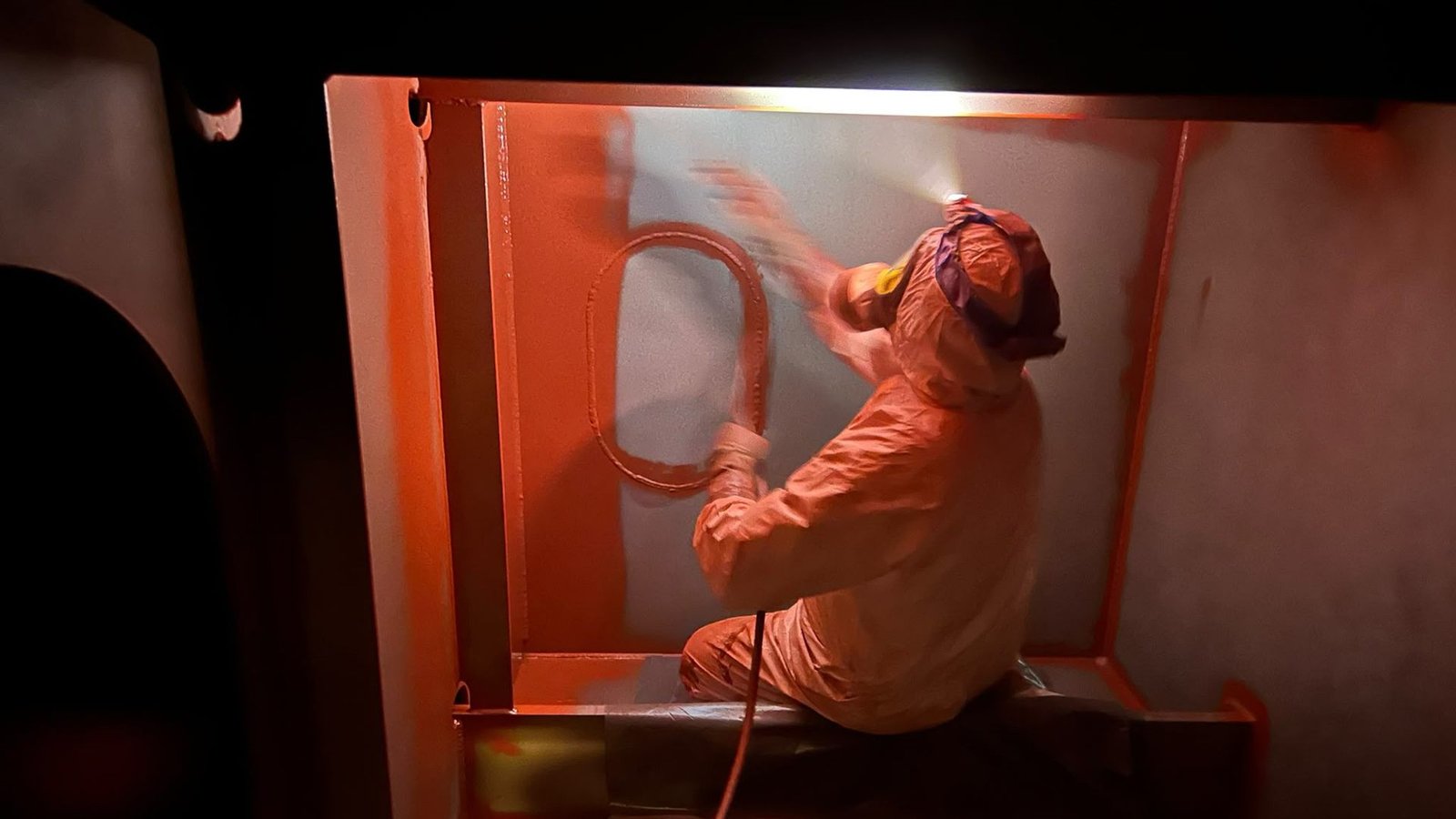 Technician in a protective suit applying spray coating inside a steel structure compartment.